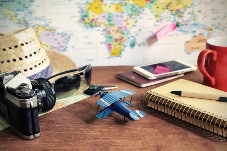 Mise en scène photographique avec divers objets représentant le voyage, l'aventure et l'histoire : un appareil photo, un chapeau de paille, des lunettes de soleil, un passeport, un avion miniature, une carte du monde, un bloc-notes avec stylo, un mug et un smartphone